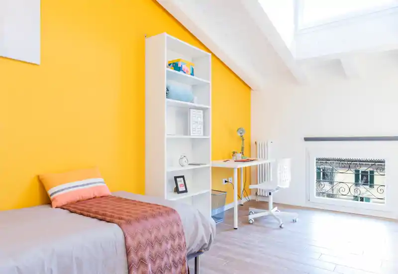 Habitación individual moderna y luminosa con pared amarilla, cama arreglada, estantería abierta y escritorio junto a una amplia ventana en el techo.