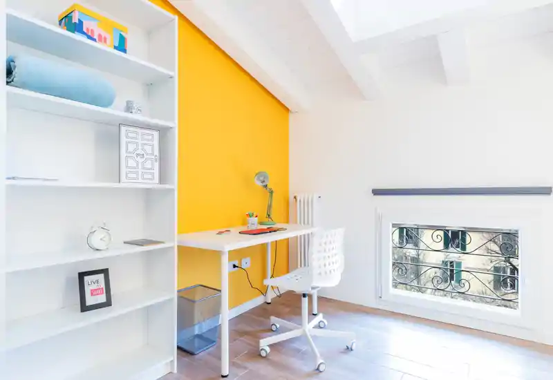 Bright, modern small bedroom corner styled as a study space with a white desk, ergonomic chair, open shelving and a bold yellow accent wall providing a fresh, airy feel.
