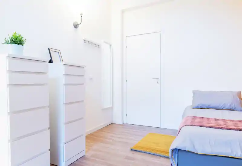 Bright, minimalist bedroom corner with a neatly made bed, soft pillows and a light throw; white storage drawers and a full-length mirror complete the airy layout.