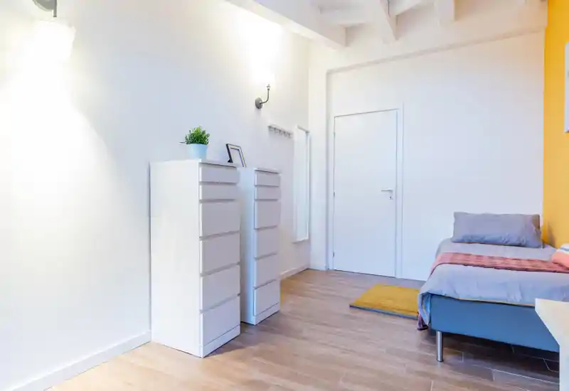 Bright, minimalist single bedroom with a neatly made bed against a warm accent wall, two slim white drawers and wooden flooring. The space is clean, well-lit and styled for a cozy single stay.