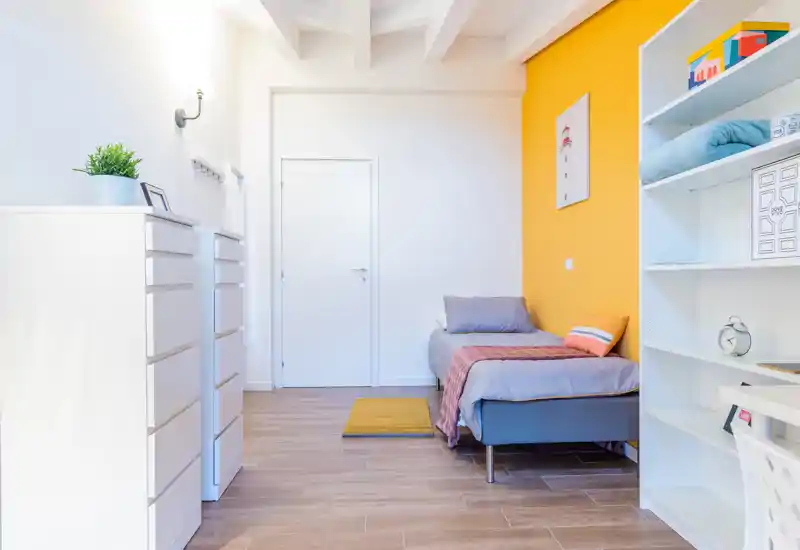 Camera singola luminosa e moderna con letto accogliente contro una parete gialla, pavimento in legno e scaffalatura pratica — perfetta per uno studente o un single.