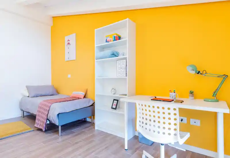 Dormitorio individual moderno y luminoso con cama bien hecha, estantería blanca y escritorio — ideal para estudiantes o un inquilino.