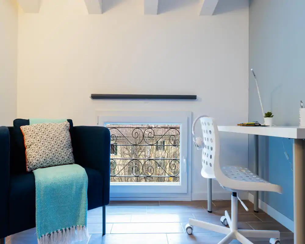 Area living moderna e luminosa con una poltrona blu scuro con coperta turchese, una scrivania e una sedia ergonomica vicino a una finestra bassa che lascia entrare luce naturale.