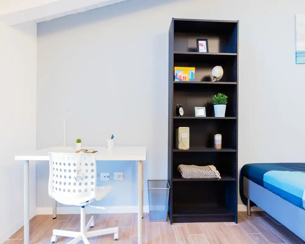 Angolo camera luminoso e moderno con scrivania, libreria e letto singolo parzialmente visibile — linee pulite e arredo minimal adatto a studenti o affitti brevi.