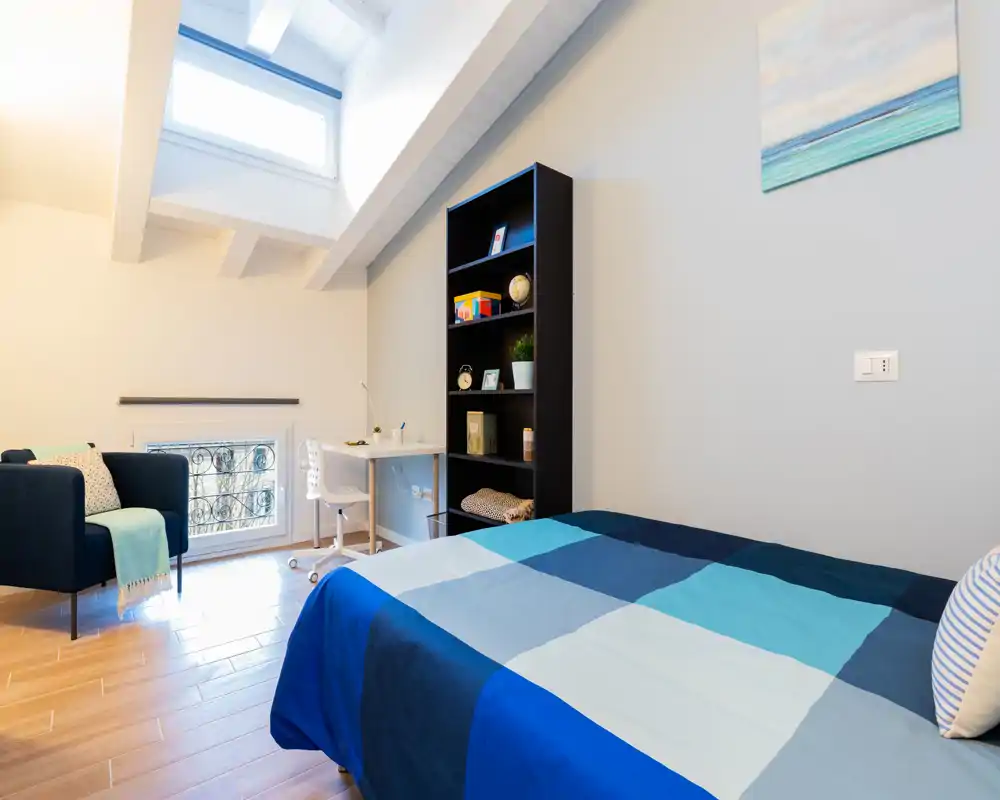 Camera da letto moderna e luminosa con coperta a quadri blu, cuscino, libreria e una scrivania; poltrona e finestra a soffitto rendono l'ambiente accogliente e ben illuminato.