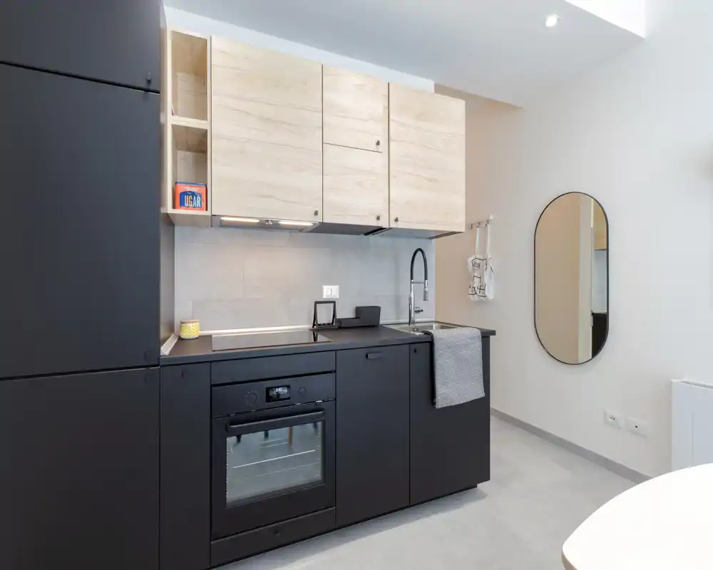 Compact modern kitchenette with matte black lower cabinets, light wood wall cupboards and integrated oven. Clean lines, a minimalist faucet and tasteful accents create a bright, contemporary cooking area ideal for small apartments.