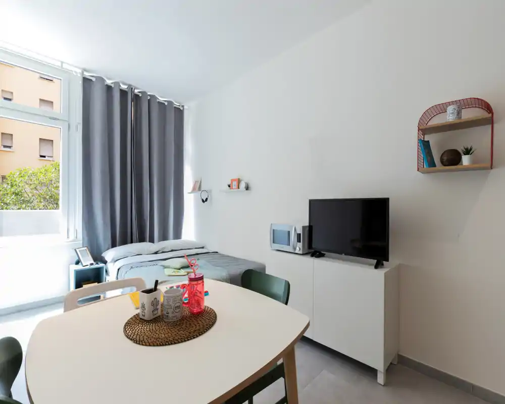 Bright, compact bedroom-studio with a double bed by a large window, simple modern furnishings and a small dining table — ideal for short stays.