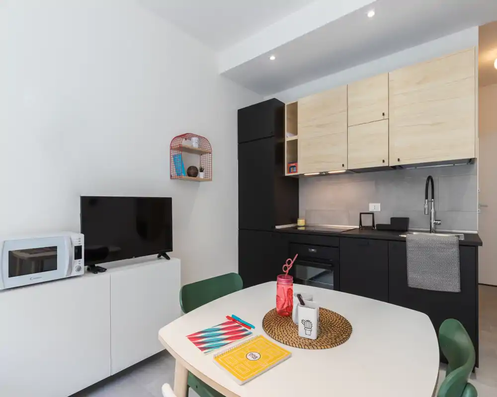Compact modern open kitchen with integrated oven, sink and upper wood cabinets; small dining table set for two adds a cozy, functional feel.