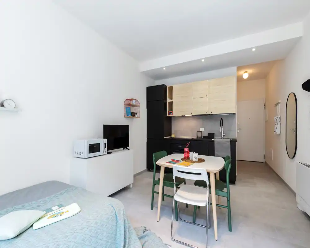 Bright, modern studio bedroom with an integrated kitchenette and a small dining table; light wood cabinets and neutral tones create a clean, functional space ideal for a single occupant or couple.