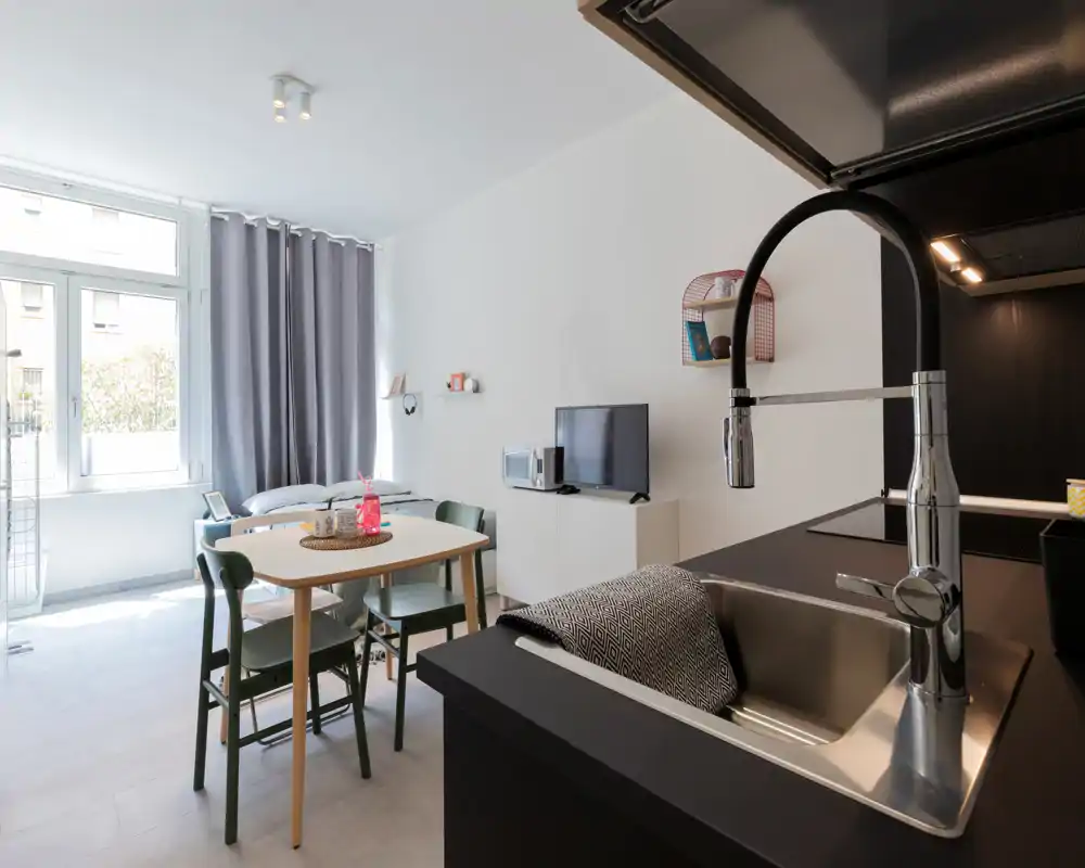 Bright open-plan kitchenette with a modern sink in the foreground, a small dining table and a daybed by the window — ideal for a compact studio apartment.