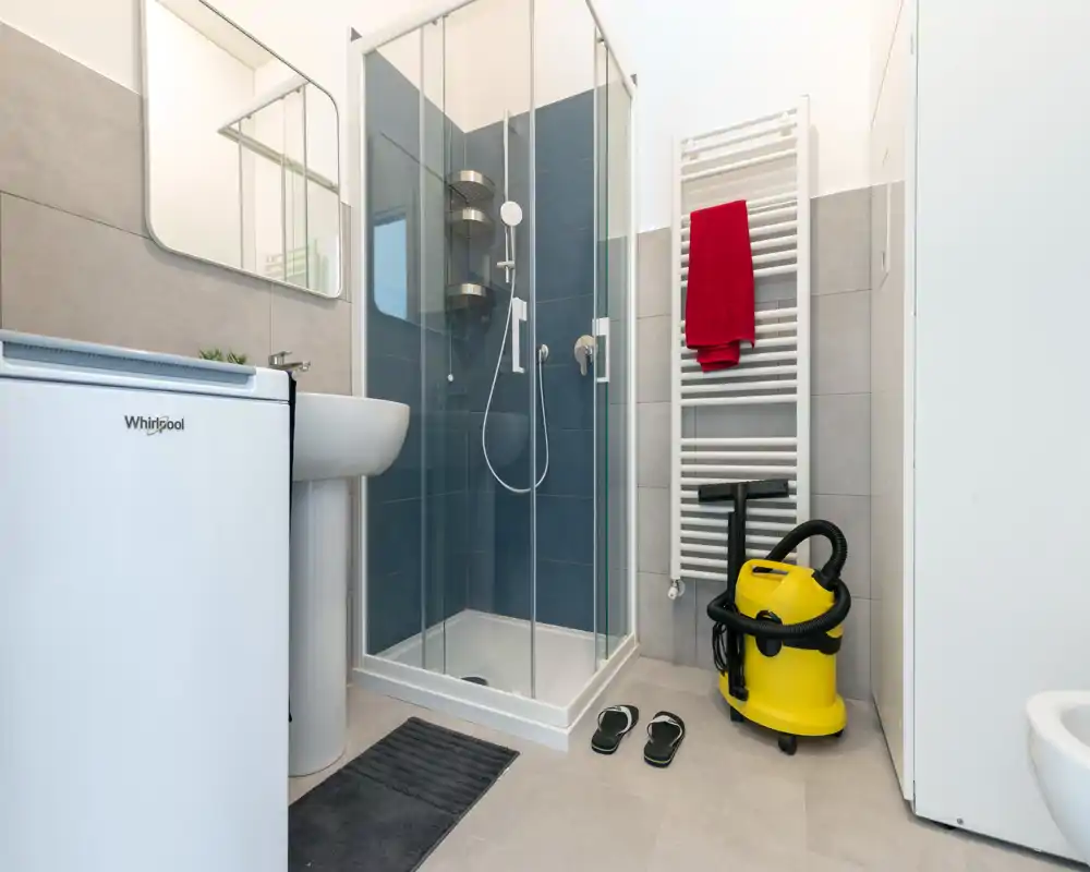 Modern compact bathroom with a glass shower enclosure, pedestal sink, heated towel rail with a red towel, and light grey tiles. The space is clean, well-lit and functional, ideal for a rental listing.