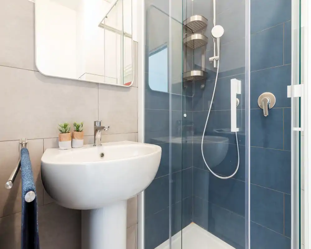 Compact modern bathroom with a pedestal sink and a glass shower enclosure with blue tiles; bright, clean and simply styled.