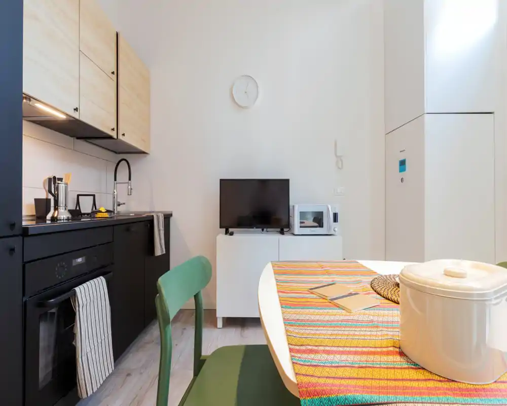 Rincón de cocina moderno y compacto con muebles bajos negros, armarios altos de madera, horno integrado y fregadero; pequeña mesa con camino de mesa colorido y sillas verdes, TV y microondas en un mueble lateral — brillante y bien decorado.