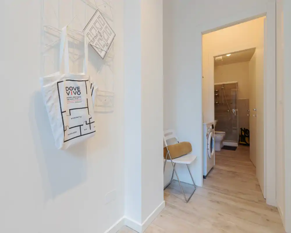 Entrada/pasillo luminosa y minimalista con suelo de madera clara, una silla plegable y un colgador con un bolso; a través de la puerta abierta se ven la zona de lavandería y el baño.