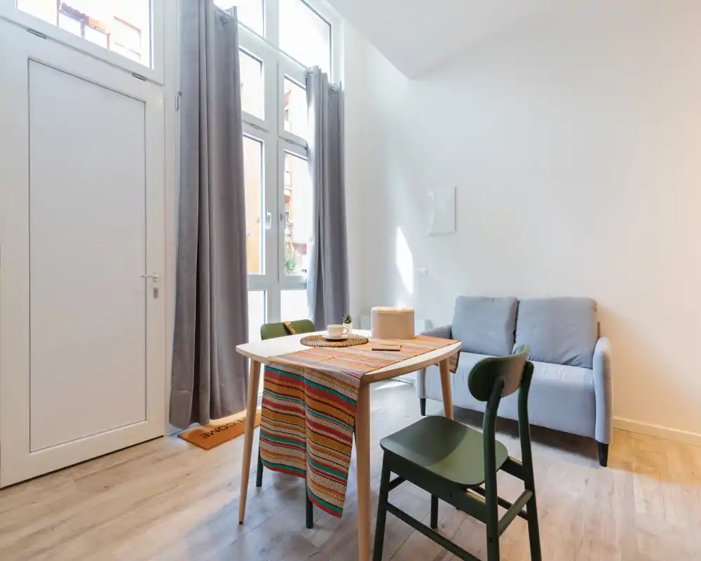 Zona de estar compacta y luminosa con sofá pequeño, mesa de comedor y ventanas altas que dejan entrar abundante luz natural — ideal para un apartamento urbano y acogedor.