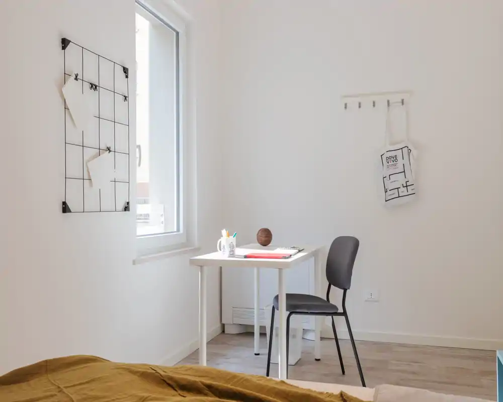 Rincón de dormitorio luminoso y minimalista con una pequeña zona de trabajo junto a la ventana. La cama se ve parcialmente en primer plano y hay un escritorio ordenado para estudiar o teletrabajar.