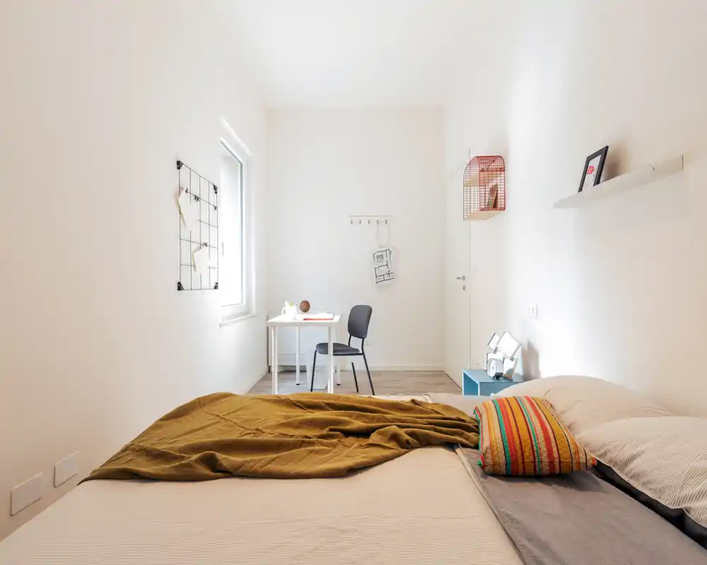 Dormitorio luminoso y minimalista con una cama grande en primer plano, cojín colorido y un escritorio junto a la ventana. La habitación es amplia y bien iluminada, ideal para una persona o una pareja que busque un espacio moderno y tranquilo.