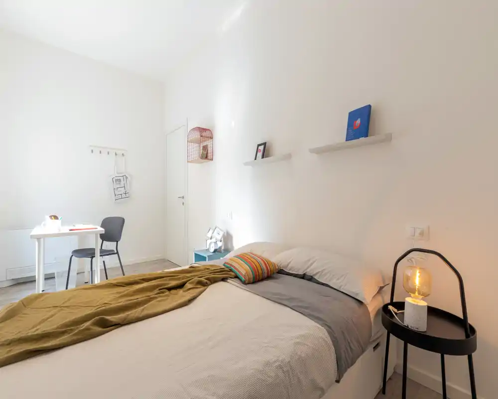 Dormitorio luminoso y moderno con cama doble cómoda, mesita con lámpara cálida y un pequeño escritorio junto a la ventana — ideal para una estancia acogedora y minimalista.