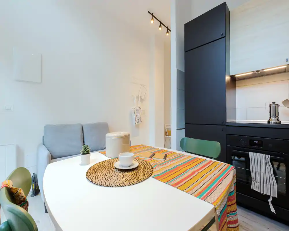 Rincón de cocina moderno y luminoso con mesa para dos, camino de mesa colorido y mantel, electrodomésticos negros empotrados y asientos acogedores: perfecto para un piso compacto.