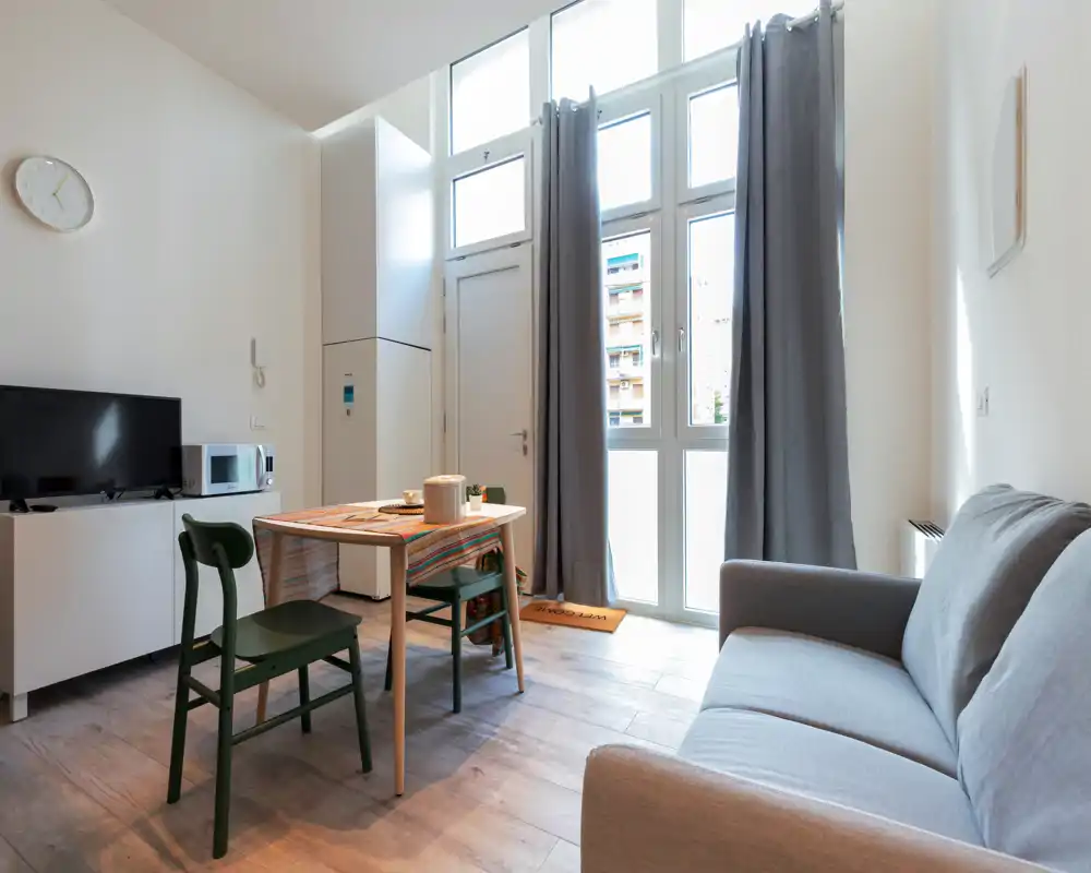 Sala de estar moderna y luminosa con una mesa de comedor compacta, dos sillas y un sofá cómodo junto a grandes ventanas que aportan luz natural.