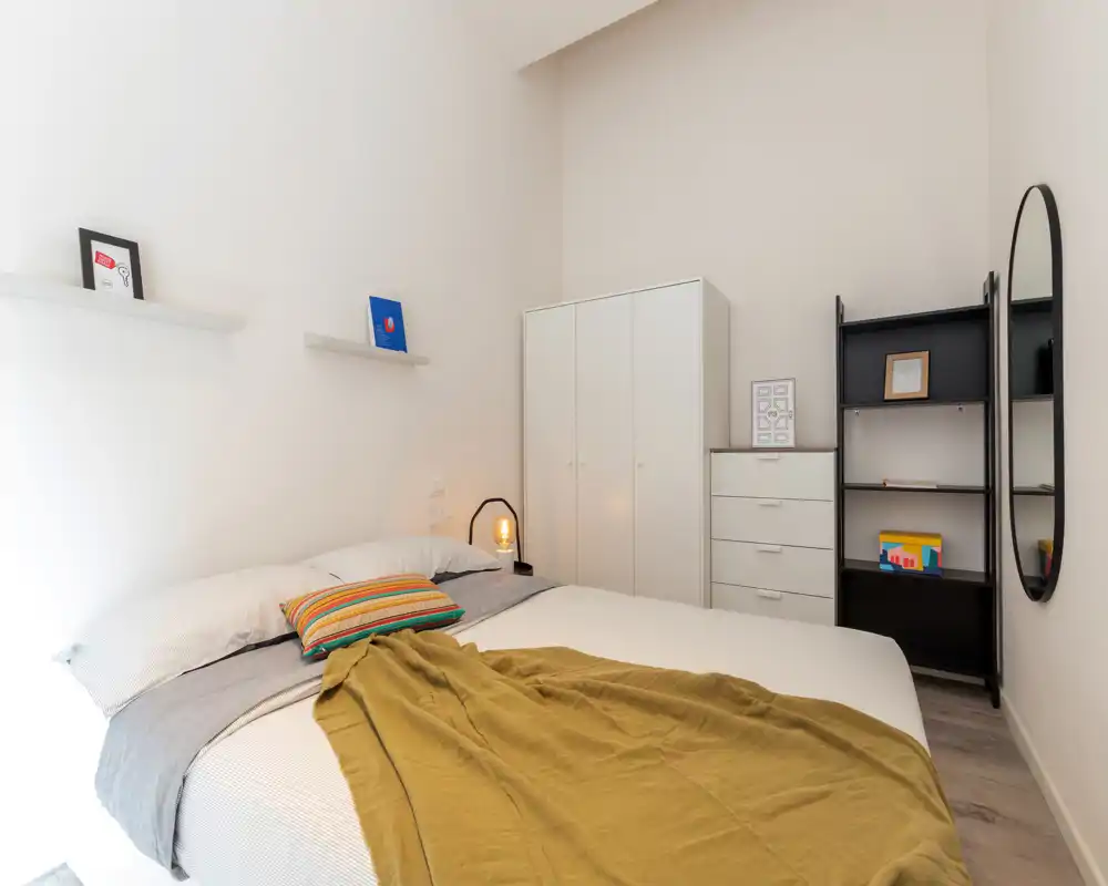 Dormitorio luminoso y minimalista con cama doble bien hecha, manta texturizada, lámpara de mesita y armarios sencillos — líneas limpias y luz natural que crean un ambiente tranquilo.