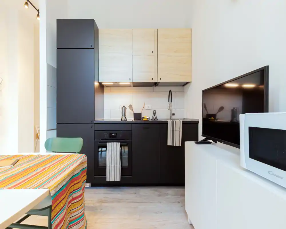 Cocina compacta y contemporánea con muebles inferiores negros mate, armarios superiores en madera clara, horno empotrado y pequeño fregadero: diseño luminoso y funcional para un piso urbano.