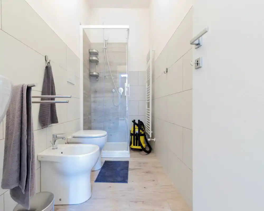 Baño moderno y luminoso con ducha de cristal, radiador toallero en pared, bidé y WC sobre suelo efecto madera. Azulejos neutros y distribución ordenada que aportan un espacio limpio y funcional.