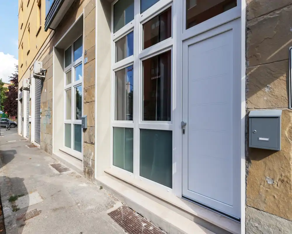 Entrada a la calle con puerta blanca moderna y ventanas inferiores esmeriladas; buzón y fachada de piedra visibles, adecuada para mostrar el acceso del edificio.