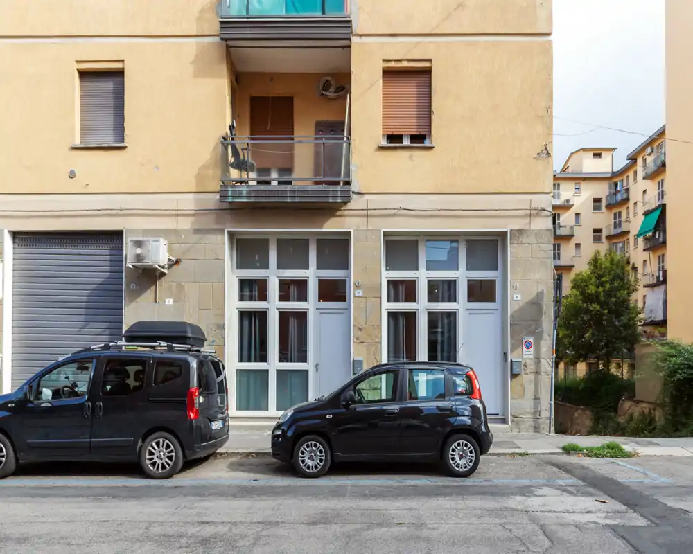 Fachada a nivel de calle de un edificio residencial con dos coches aparcados, puertas y ventanas en planta baja y un pequeño balcón arriba.