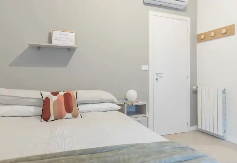 Dormitorio moderno y luminoso con cama doble bien hecha, cojín decorativo y mesita con lámpara — paleta neutra y buena luz.