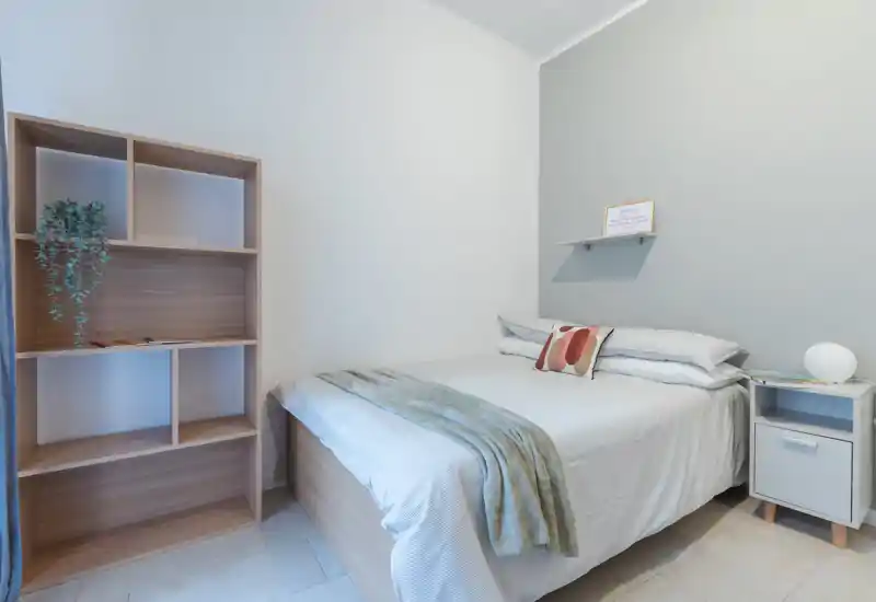 Dormitorio luminoso y minimalista con cama doble bien preparada, ropa de cama suave, mesita con lámpara y estantería para almacenaje — ideal para una estancia acogedora y moderna.
