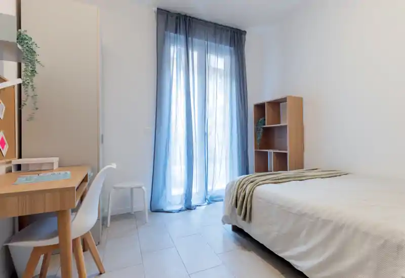 Dormitorio luminoso y sencillo con cama individual, escritorio y estantería; cortina traslúcida deja pasar luz natural suave.