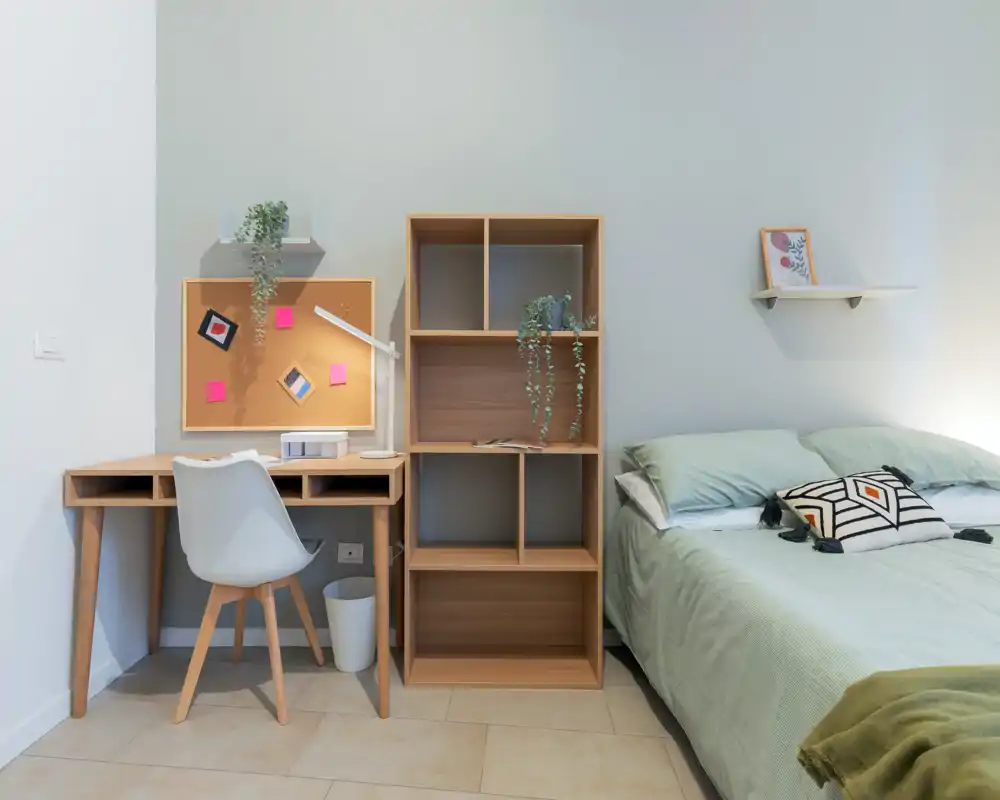 Camera da letto luminosa e ordinata con un comodo letto matrimoniale, biancheria verde chiaro, un cuscino decorativo e una libreria in legno accanto a una scrivania compatta — uno spazio accogliente e ben illuminato ideale per una persona o una coppia.