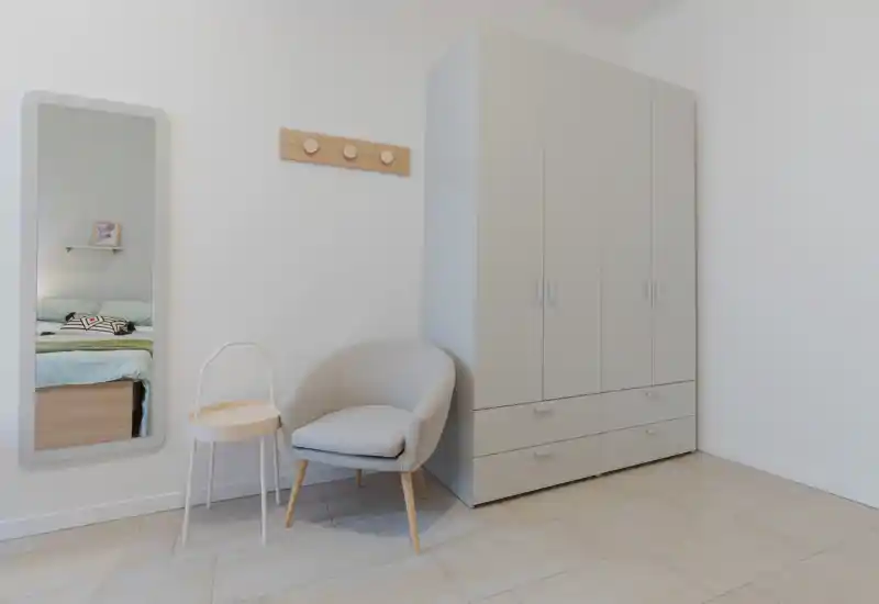 Angolo di camera da letto minimal con grande armadio, una poltroncina e uno specchio a figura intera che riflette parte del letto. Toni neutri e arredi moderni creano un’atmosfera luminosa e rilassante, ideale per annunci.