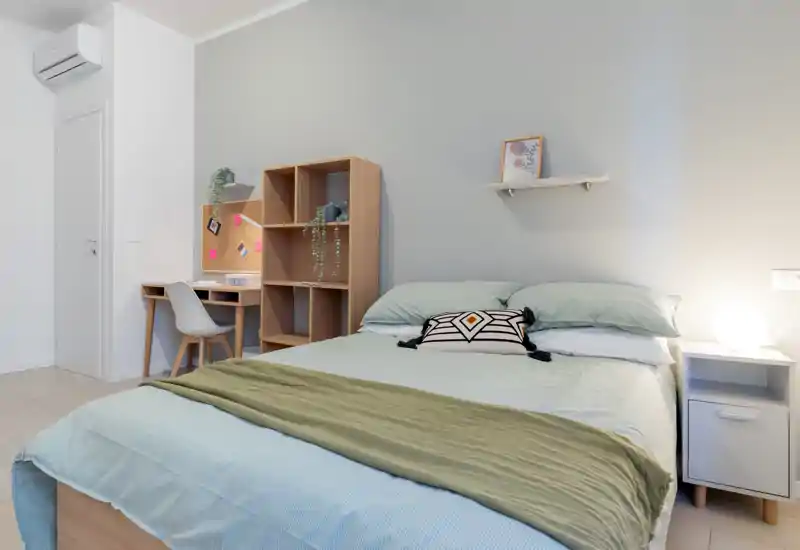 Camera da letto moderna e luminosa con letto matrimoniale ben rifatto, comodino con lampada e un piccolo angolo studio con scaffali — perfetta per studenti o professionisti.