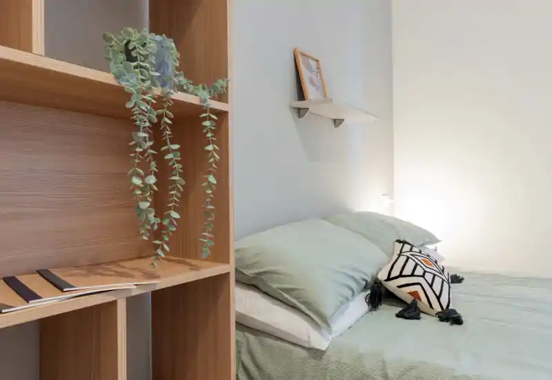 Camera da letto luminosa e minimal con letto ben rifatto in lenzuola verde salvia, cuscino decorativo e luce soffusa laterale; scaffalatura in legno con pianta pendente che aggiunge calore naturale.