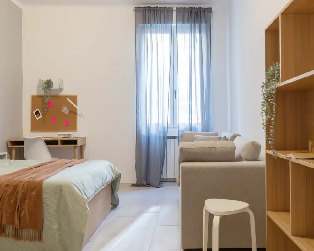 Camera da letto luminosa e minimal con letto comodo, una piccola scrivania e un'area salotto accogliente vicino alla finestra, ideale per una persona o uno studente.