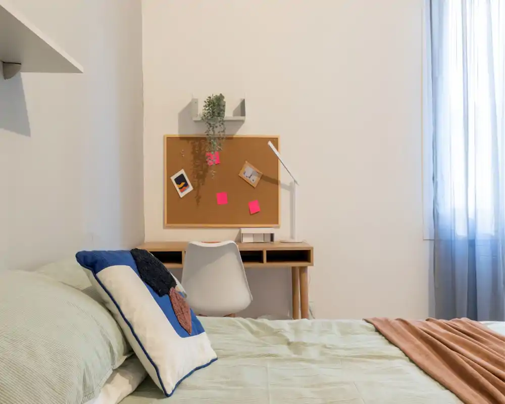 Camera da letto ben arredata con un letto accogliente in primo piano, cuscini decorativi e una scrivania compatta con una bacheca al muro; luce naturale morbida filtra dalla tenda leggera.
