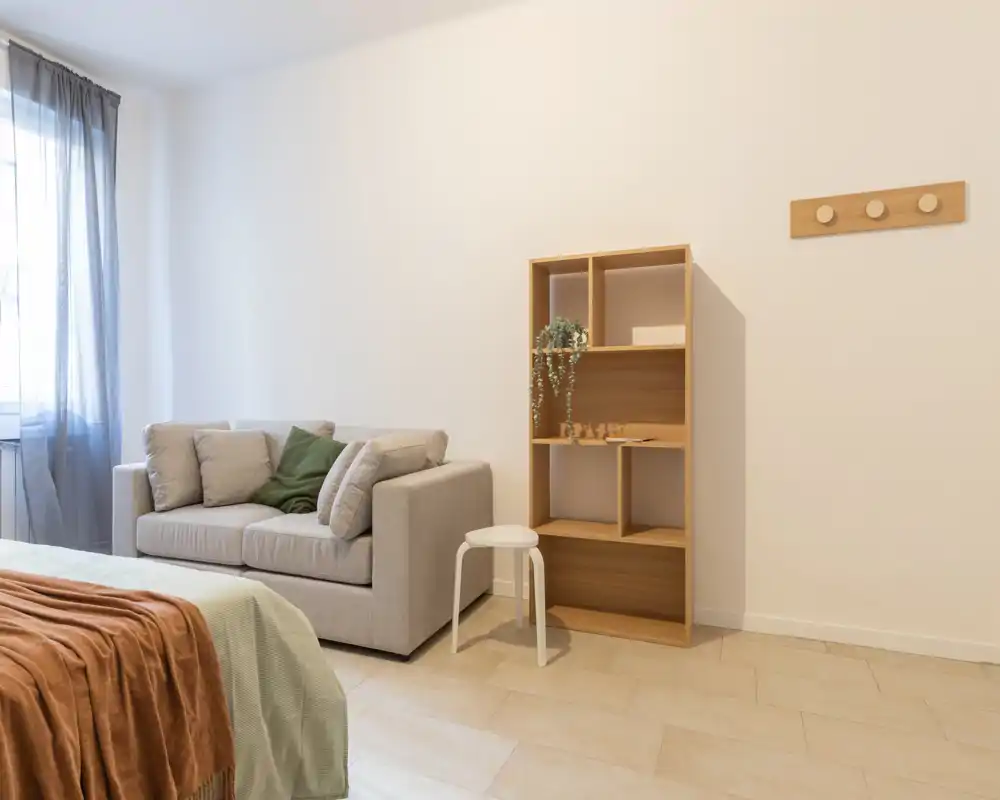 Zona notte ariosa e minimal con letto in primo piano, un piccolo divano e luce naturale da una finestra grande che crea un interno tranquillo dai toni neutri.