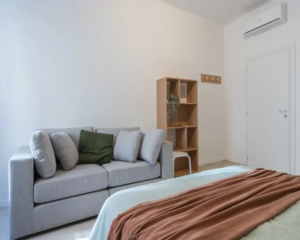 Camera da letto moderna e luminosa con letto in primo piano, un piccolo divano e una scaffalatura sullo sfondo; toni neutri e tessuti morbidi creano un ambiente rilassante.