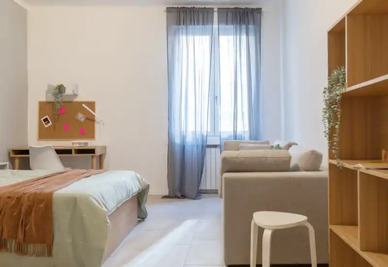 Camera da letto luminosa e minimal con letto comodo, una piccola scrivania e un'area salotto accogliente vicino alla finestra, ideale per una persona o uno studente.