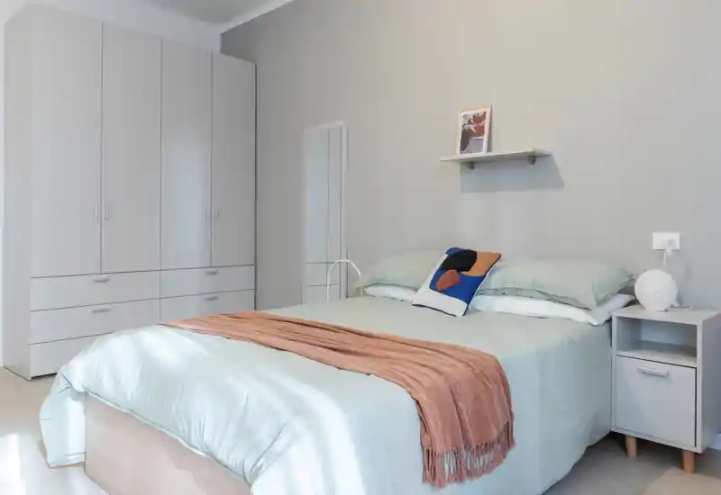 Camera da letto moderna e luminosa con un ampio letto ben rifatto, biancheria pastello, armadio e comodino — perfetta per annunci in affitto che valorizzano uno stile minimal e rilassante.