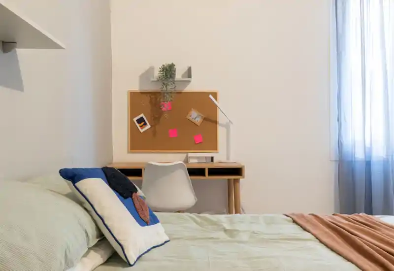 Camera da letto ben arredata con un letto accogliente in primo piano, cuscini decorativi e una scrivania compatta con una bacheca al muro; luce naturale morbida filtra dalla tenda leggera.