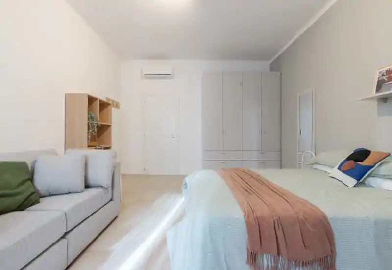 Camera da letto luminosa e moderna con un ampio letto con biancheria morbida, plaid e cuscini decorativi, un divano compatto e un armadio a muro—perfetta per un monolocale elegante.