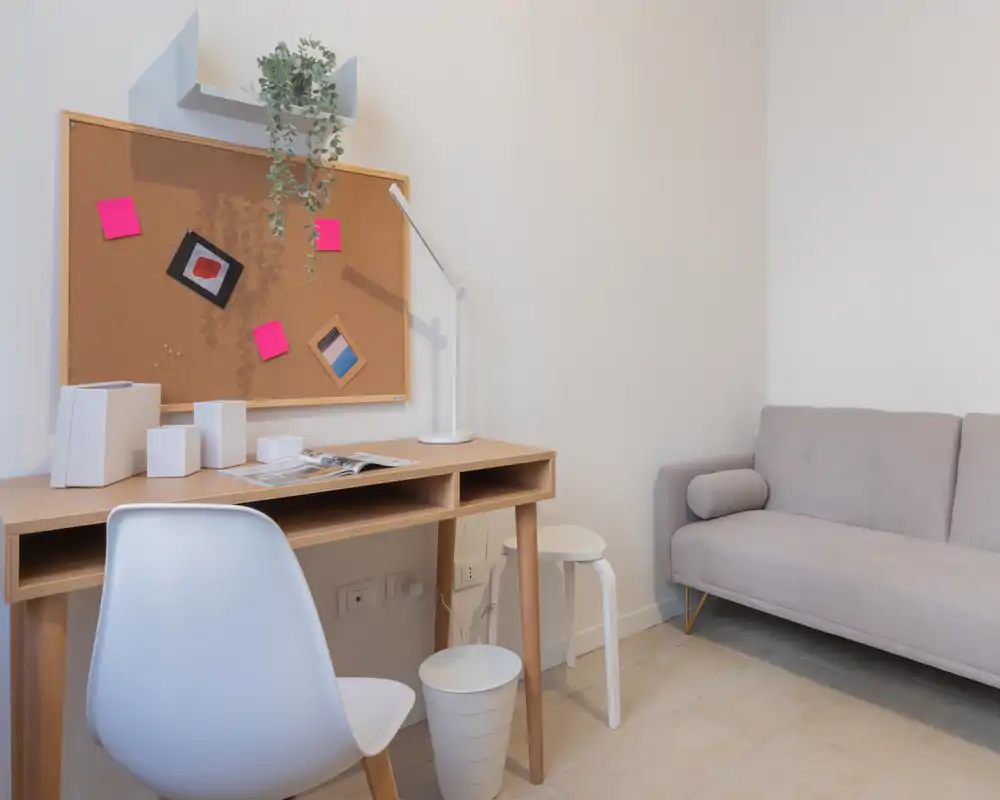 Area giorno luminosa e minimal con una scrivania in legno compatta, sedia bianca e un divano grigio chiaro. Una bacheca con appunti e una lampada da tavolo creano un angolo lavoro funzionale e ordinato accanto alla zona relax.