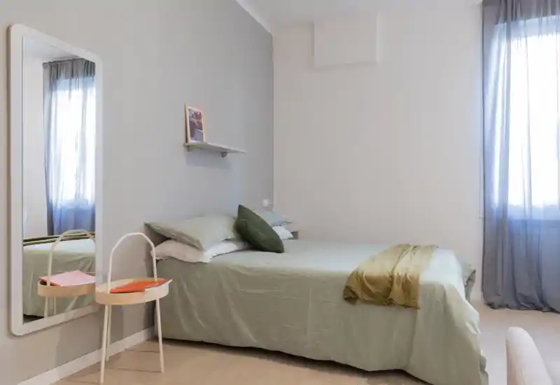 Dormitorio luminoso de estilo sencillo con cama doble, ropa de cama verde suave y luz natural por la ventana — decoración mínima y un gran espejo que aporta profundidad.
