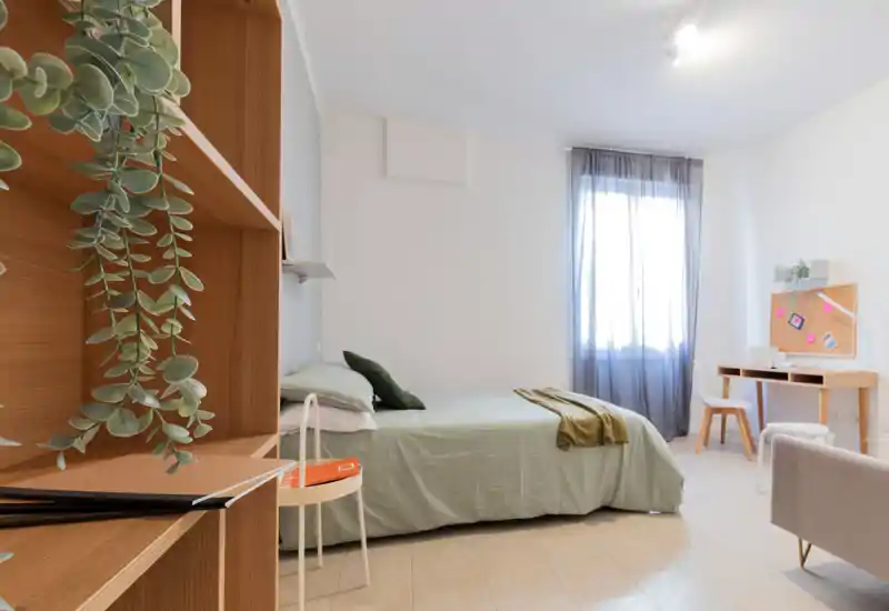 Dormitorio moderno y luminoso con cama doble bien hecha, ropa de cama verde, escritorio junto a la ventana y estanterías de madera con plantas — un espacio tranquilo y bien iluminado, ideal para viajeros o estudiantes.