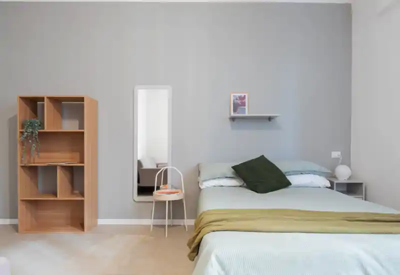 Dormitorio minimalista y luminoso con cama doble bien hecha, ropa de cama clara, cojín verde como toque y una estantería y lámpara junto a la cama; líneas limpias y tonos neutros que crean un ambiente moderno y tranquilo.