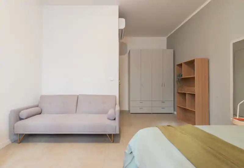 Dormitorio luminoso y minimalista con la cama visible en primer plano, un pequeño sofá junto a la pared y un armario y estantería al fondo — tonos neutros y líneas limpias.