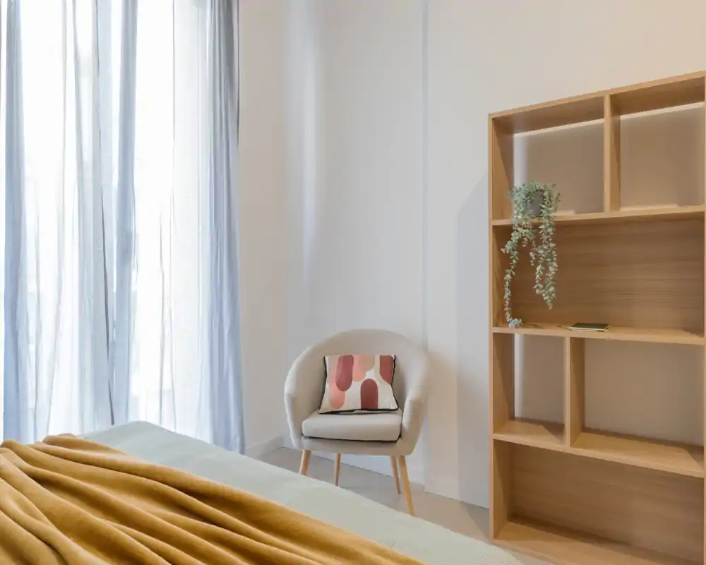 Camera da letto luminosa e minimal con letto ordinato in primo piano, poltrona accogliente con cuscino decorativo e scaffalatura in legno a vista — stile moderno e tranquillo perfetto per annunci.